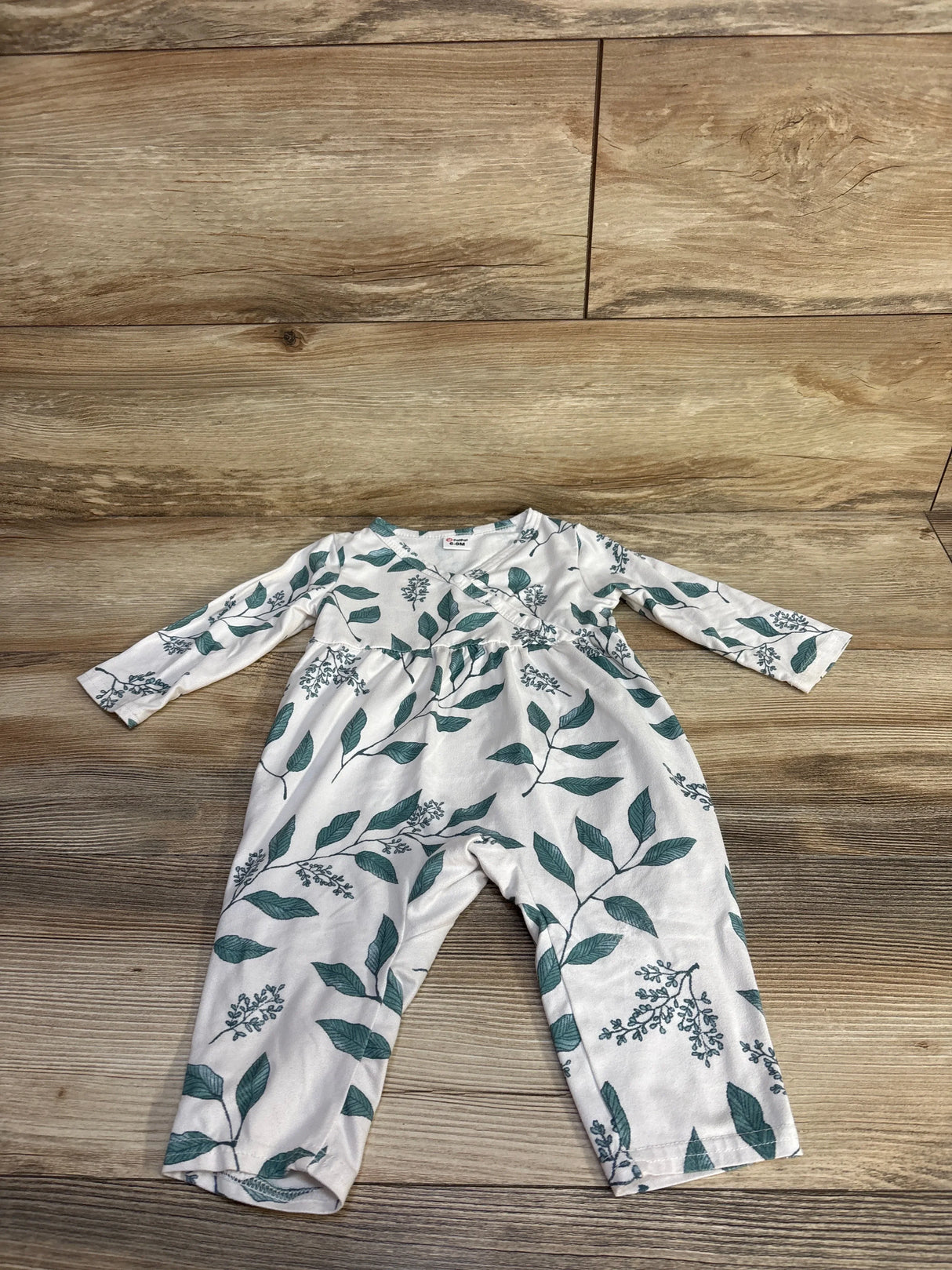 Flat lay of white baby coverall (size 6–9 months) showing green leaf print and long sleeves on wood background