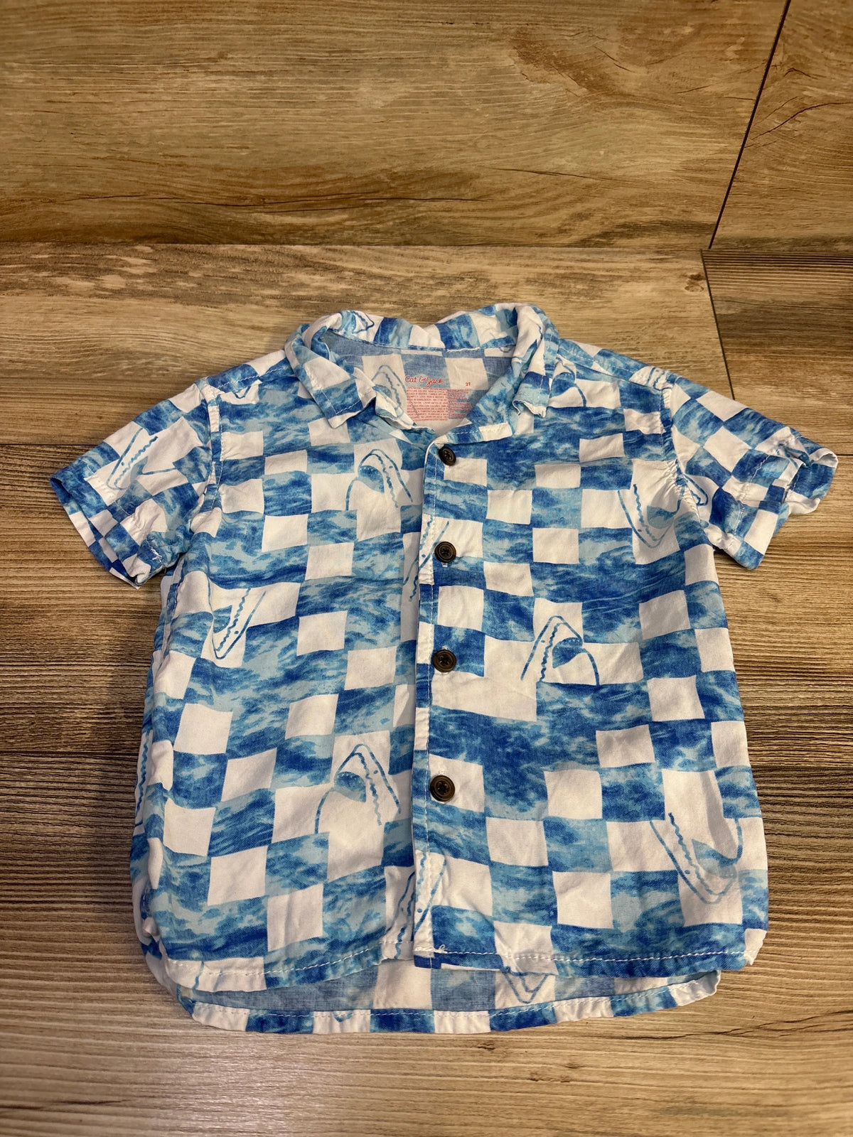 Front view of Cat and Jack 2T short-sleeve button-up shirt with blue and white checkerboard and shark pattern on wood background