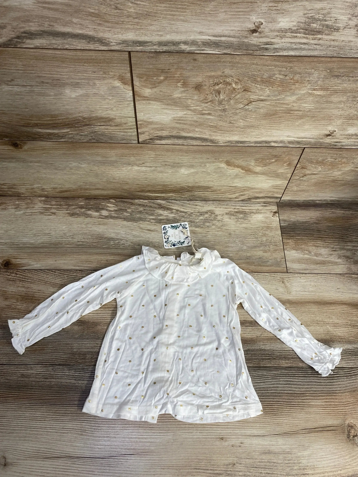 Flat lay of Ivory Chelsea & Violet long sleeve baby shirt 12m showing ruffled collar and gold polka dots on wood floor