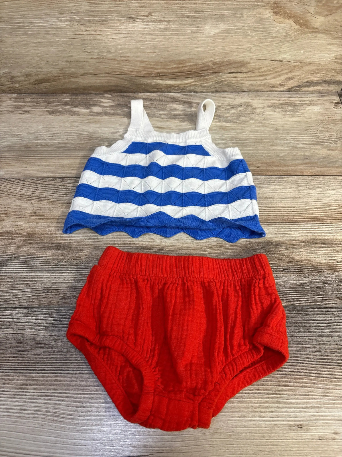 Top-down view of baby 4th of July two-piece: blue-and-white striped tank and red cotton bloomers on wood background