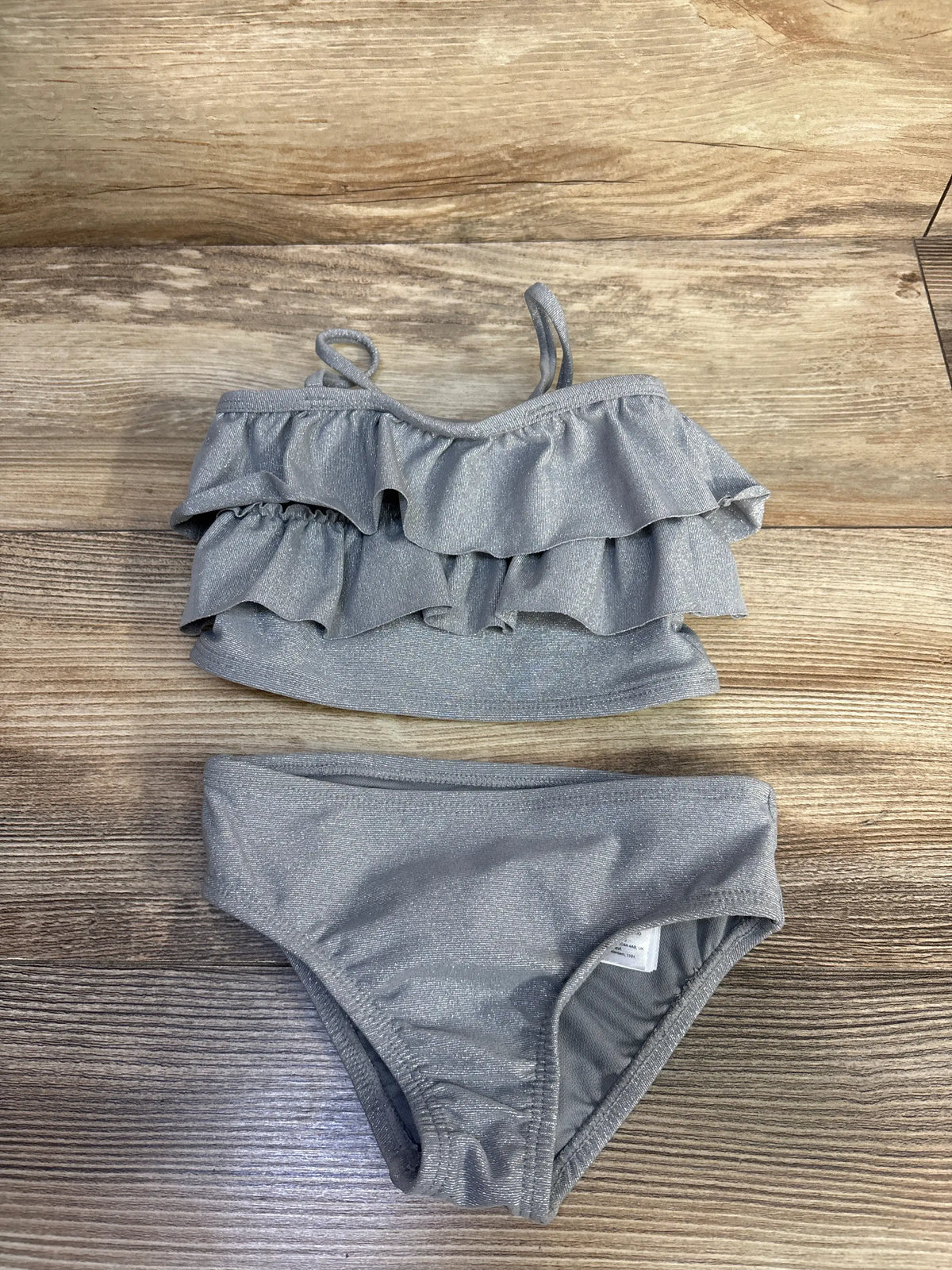 Top-down view of Baby Gap two-piece silver tankini swimsuit showcasing ruffled layered tank top and matching briefs on wooden background