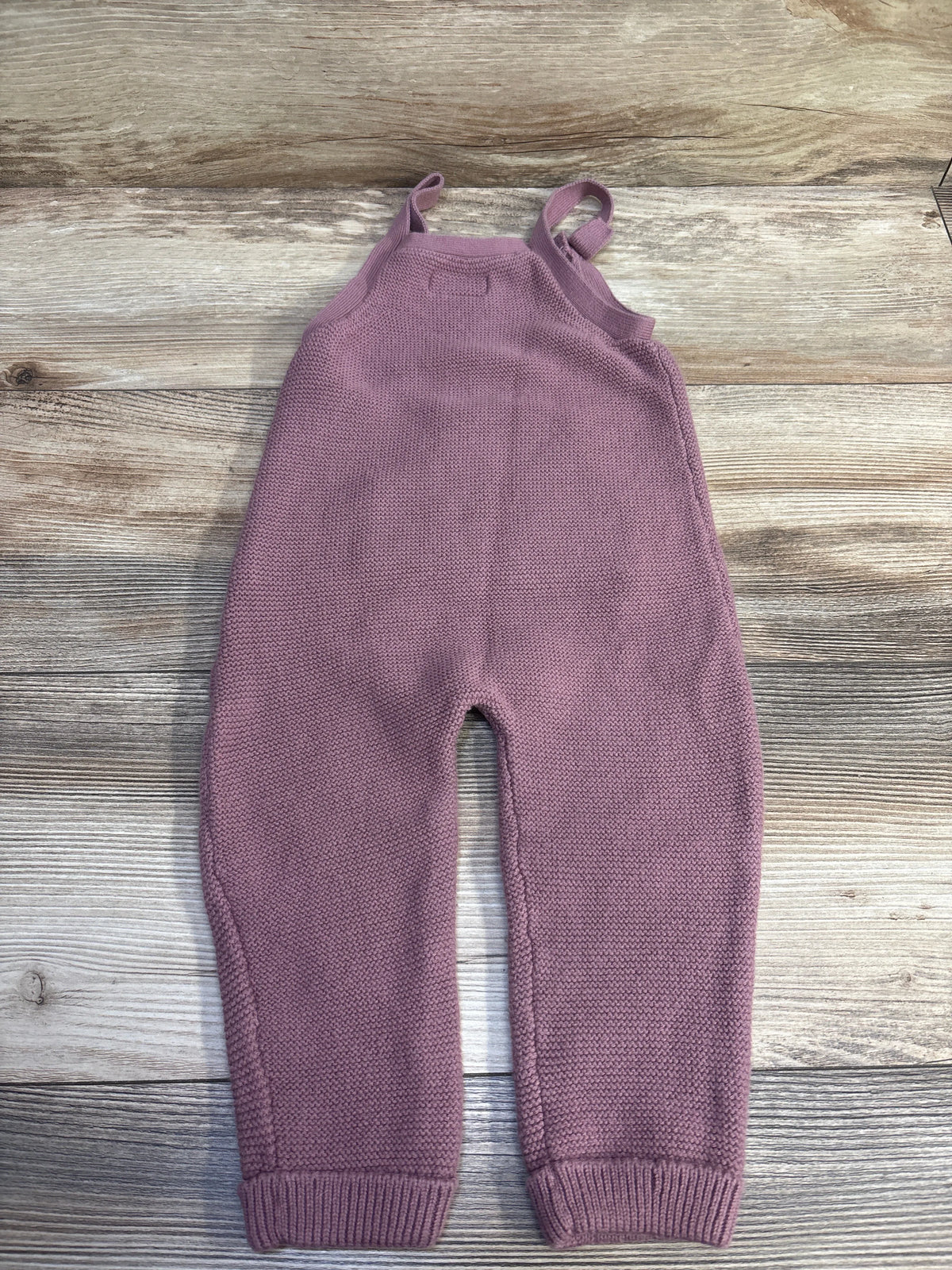 Top view of Little Planet knit overalls in mauve showcasing shoulder straps and ribbed cuffs with a wood grain background