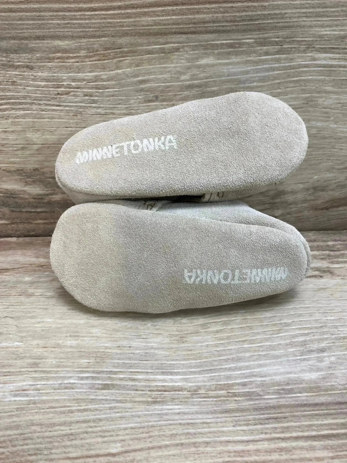 Top-down view of Minnetonka Star infant booties showcasing tan suede soles with printed Minnetonka logo on light wood background