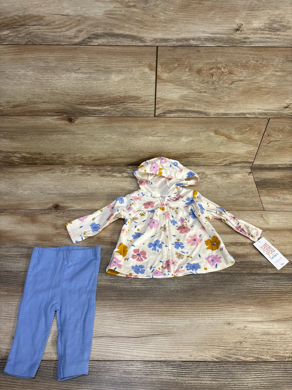 Flat-lay of 2-piece baby outfit size 3M: hooded floral zip jacket and light blue leggings on wood floor