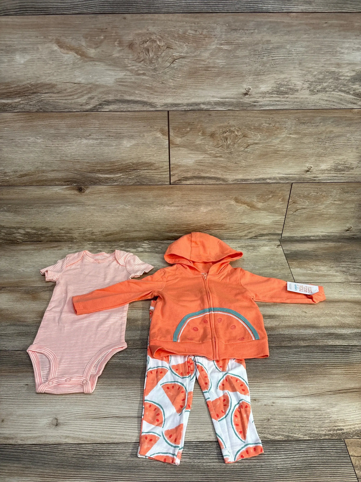 Baby clothing set with orange hooded zip-up jacket, watermelon print pants, and pink striped bodysuit on wooden floor