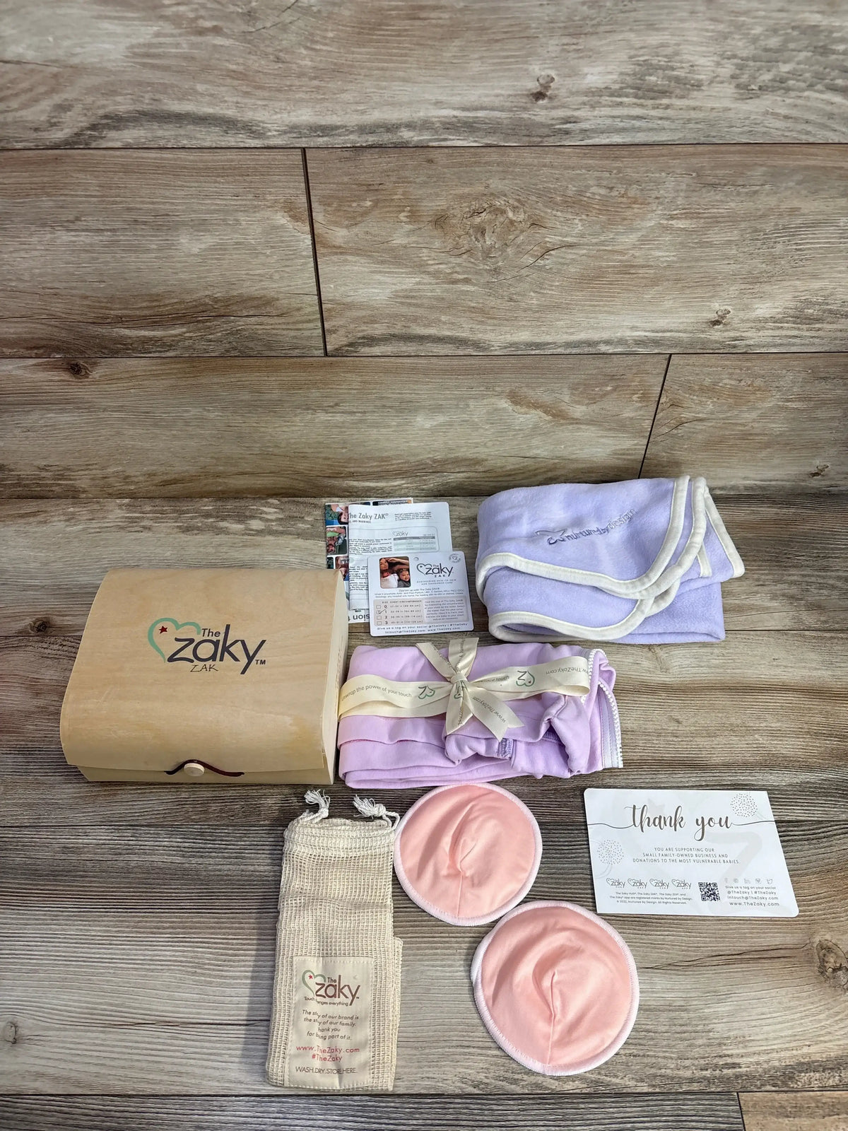Top view of The Zaky Gift Box 3pk showing wooden box, lilac swaddles, pink nursing pads, pouch and thank you card on wood