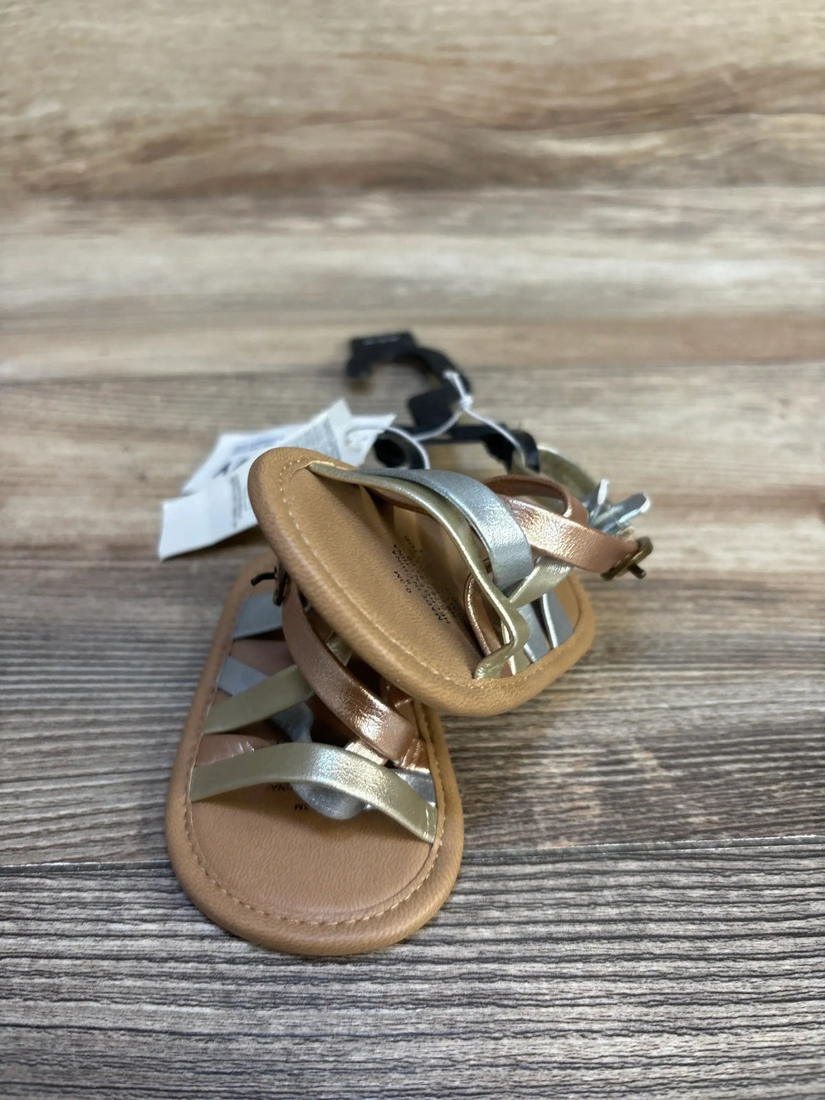 Front view of Old Navy metallic strappy baby sandals showcasing silver and rose gold crisscross straps on wood background