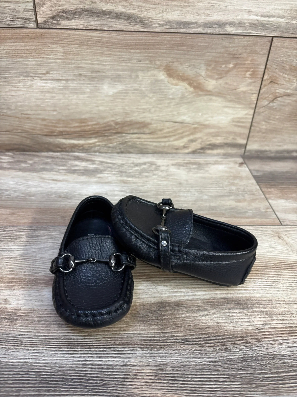 Three-quarter view of black toddler leather boat shoes showcasing silver bit and stitched moc toe on wood background