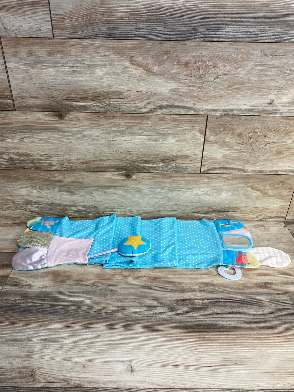 Top-down of Demdaco Mermaid activity scarf laid flat showing blue fabric, star rattle, teether, and textured panels on wood