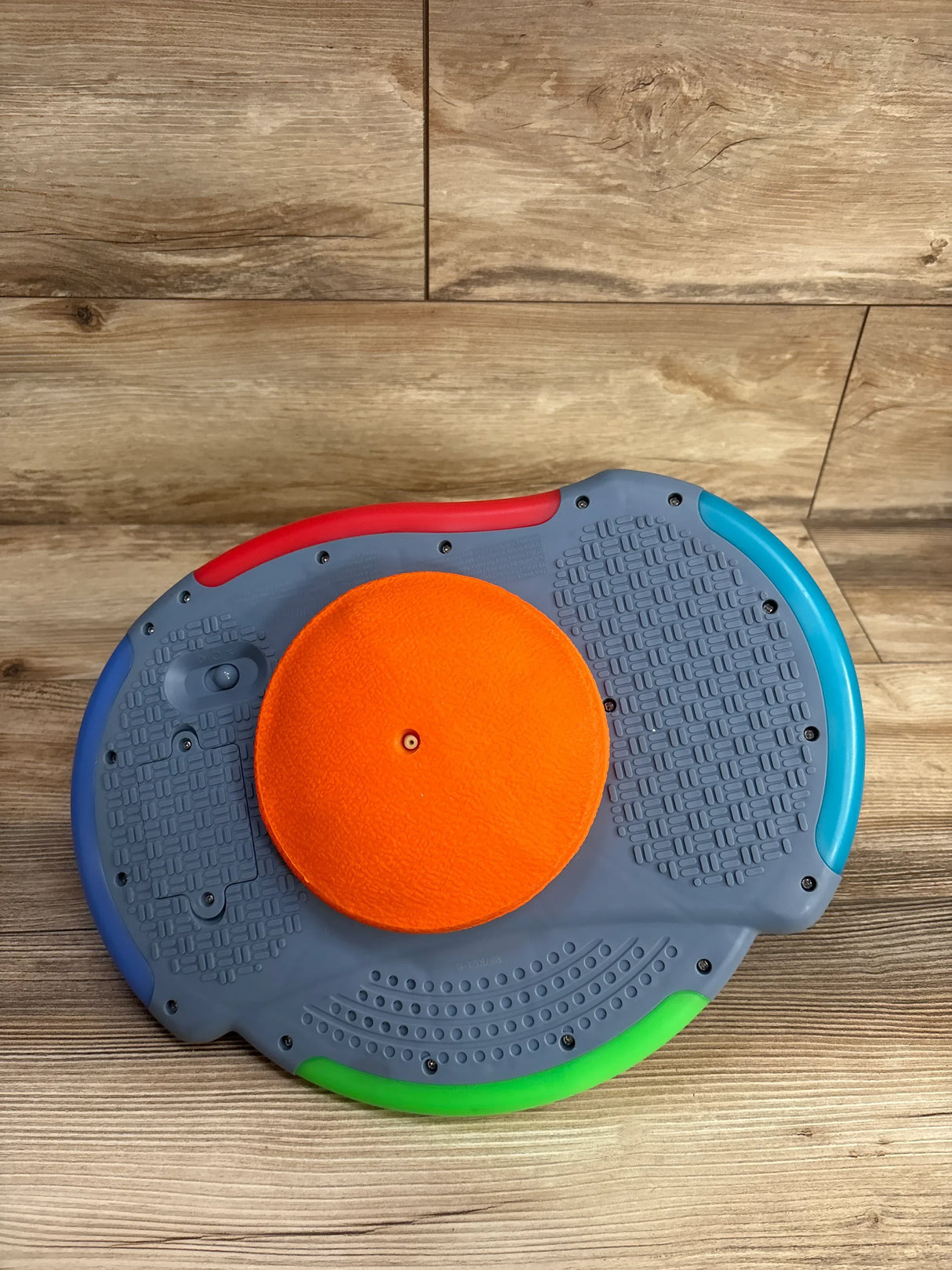 Top view of gray kids balance board showcasing orange spinner disc and red blue green colored edges on wood plank floor