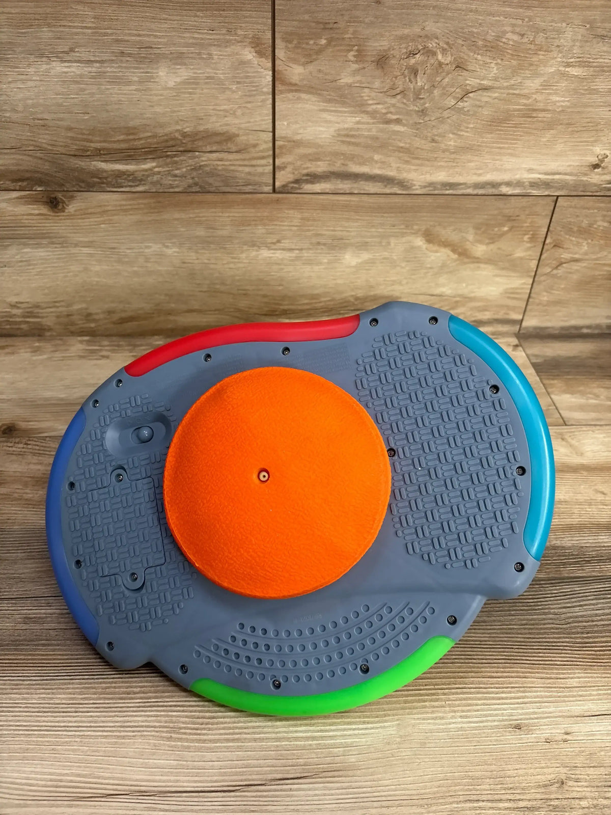 Top view of gray kids balance board showcasing orange spinner disc and red blue green colored edges on wood plank floor