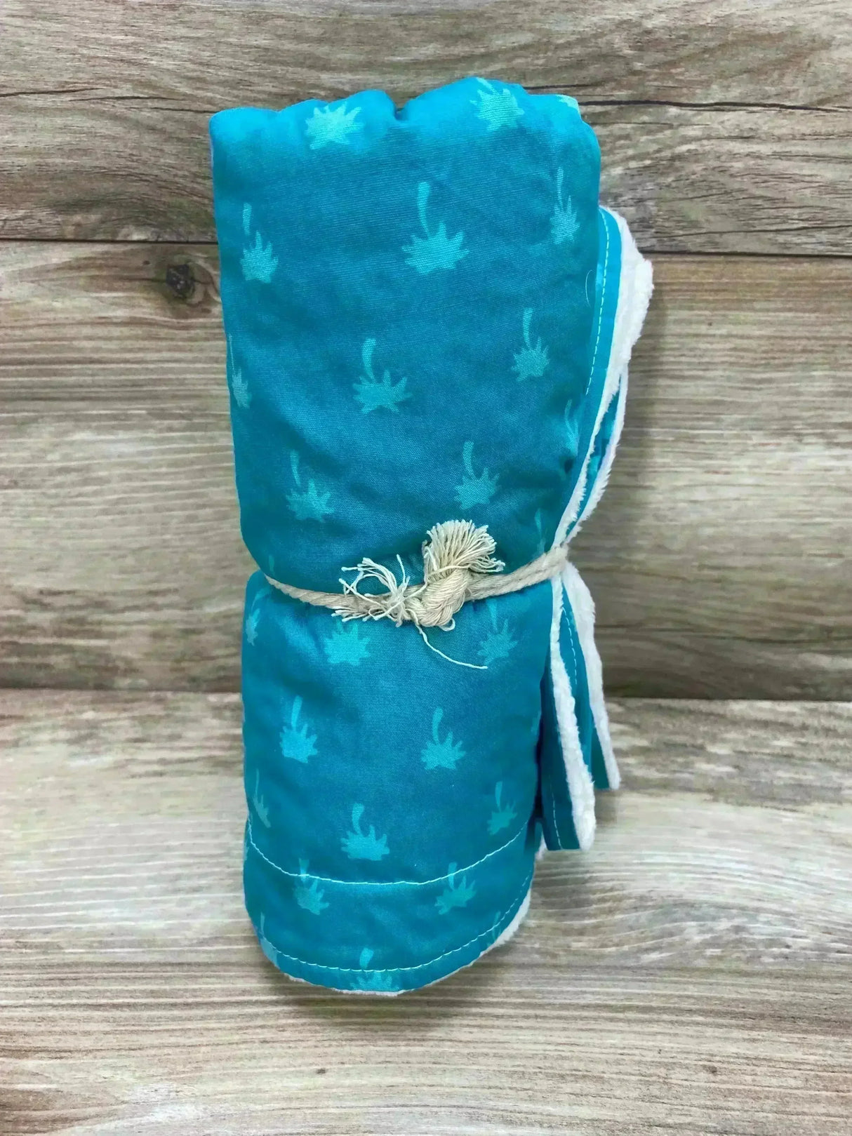 Front view of rolled Hi'ilani Hawaii teal baby blanket showing small palm-print pattern, white plush lining, and twine tie on wood plank background