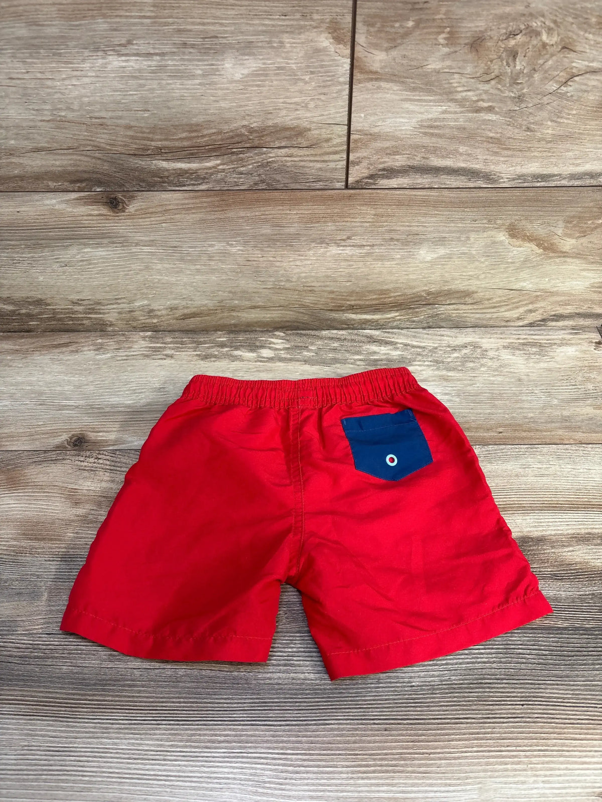 Back view flat lay of Andy & Evan toddler swim trunks in red showing elastic waistband and navy back pocket on wood background