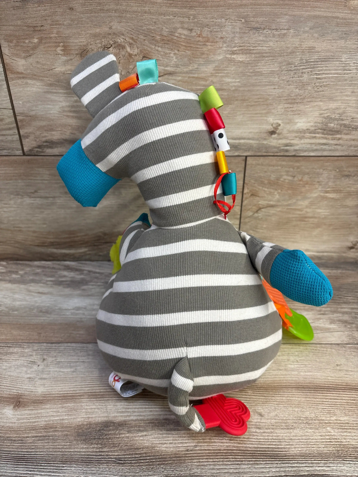 Back view of Dolce Zeddy The Zebra Activity Plush showing gray and white stripes with colorful attachments on wood