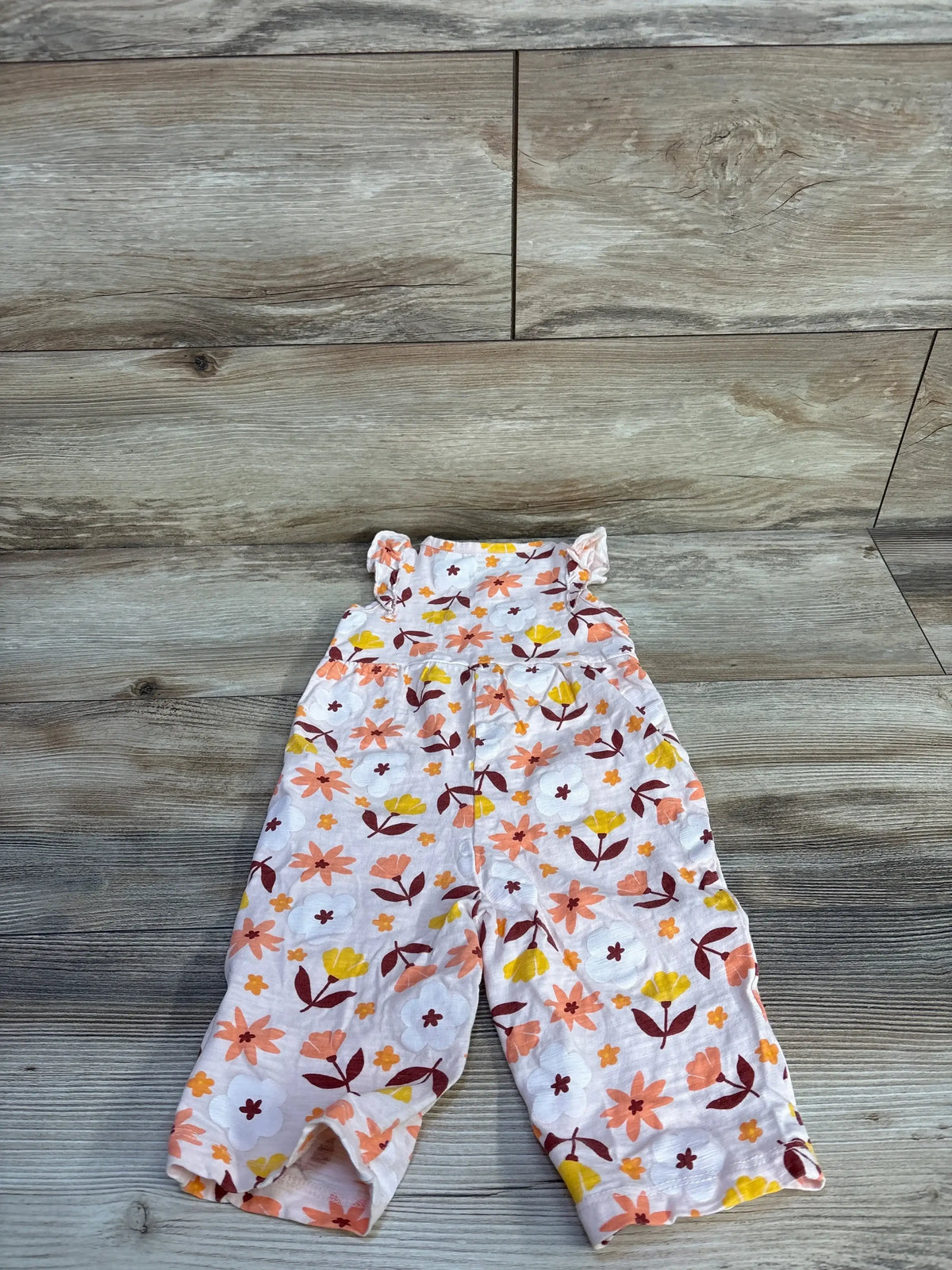 Flat lay of pink floral jumpsuit, 6-12m, showing ruffle shoulder straps and all-over flower print on wood background