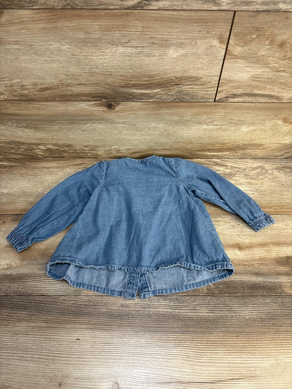 Blue denim kids long sleeve top with ruffled hem, laid flat on wood floor