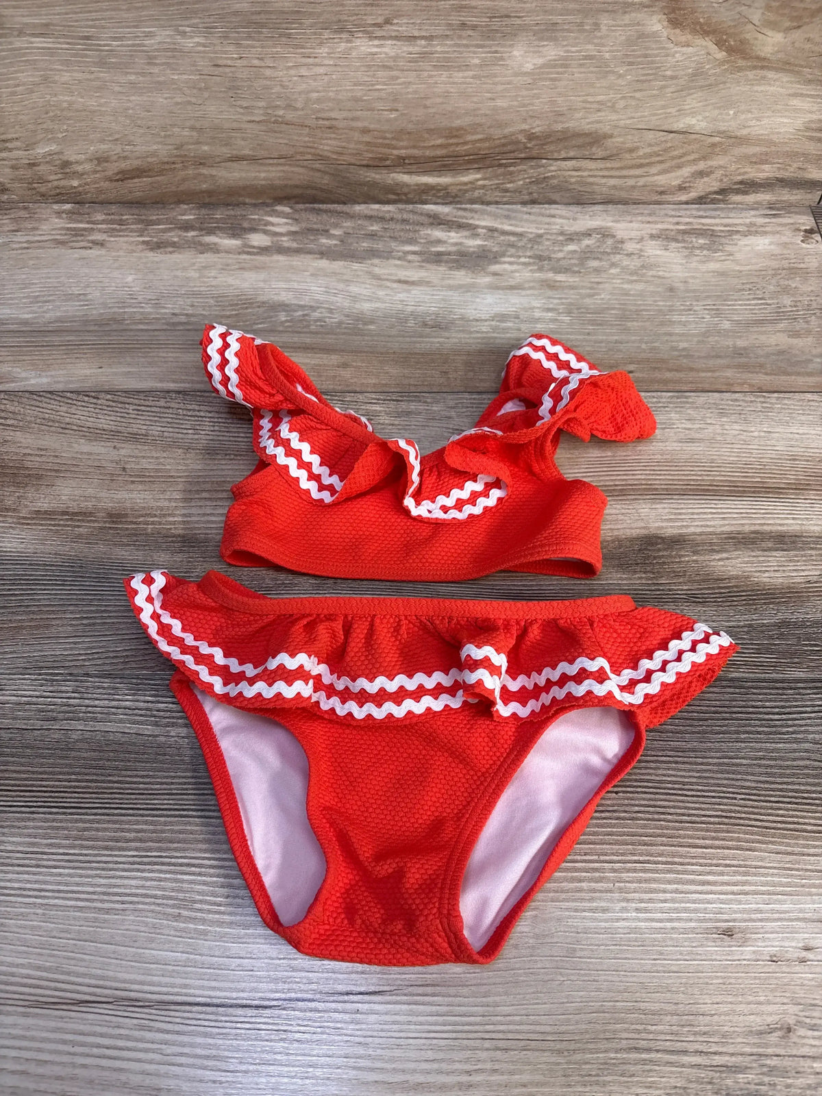 Flat lay of Janie & Jack 2pc ric-rac ruffle swimsuit showcasing red-orange textured fabric with white trim on wood plank background