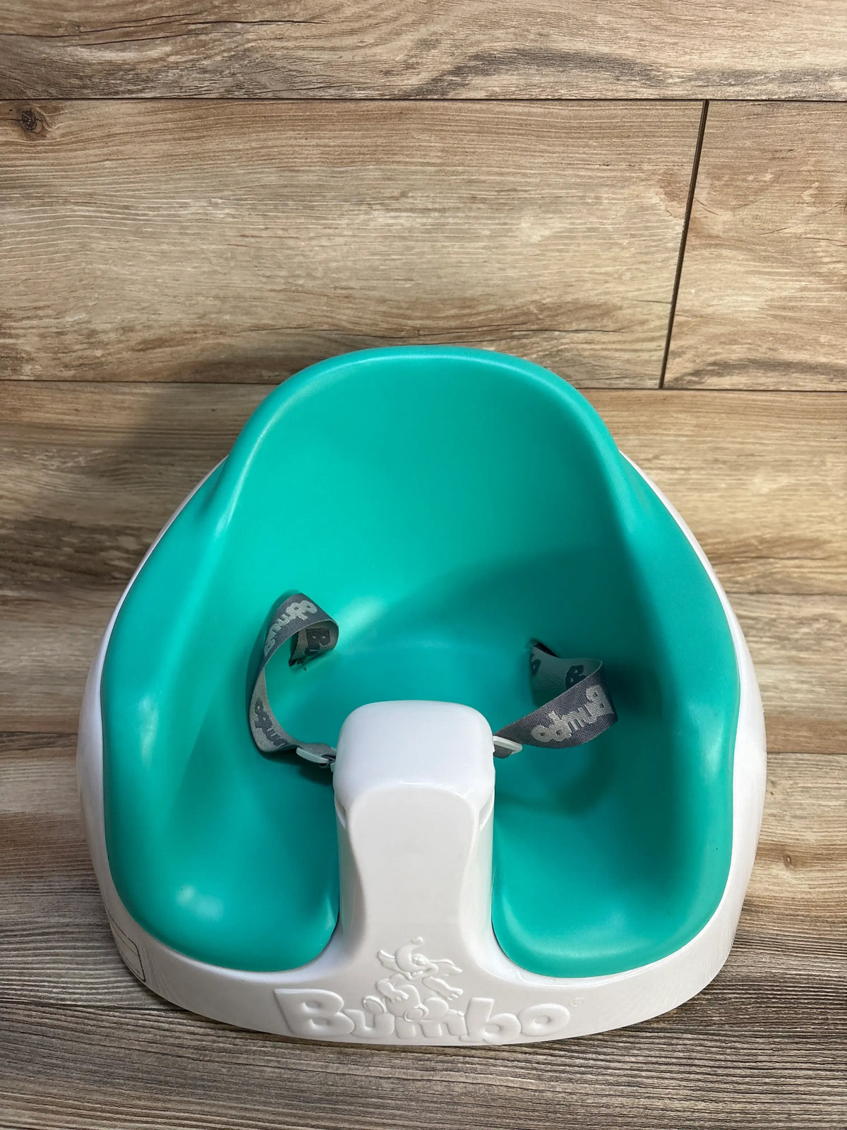 Front view of Bumbo Multi Seat in teal showcasing safety straps and a contoured seat on a wood-look floor