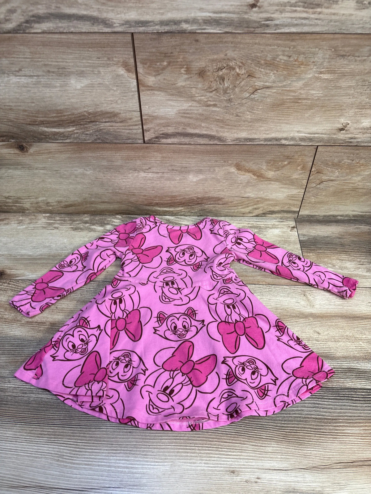 Front flat-lay of pink Minnie Mouse long-sleeve toddler dress with all-over Minnie face and bow print on wood background