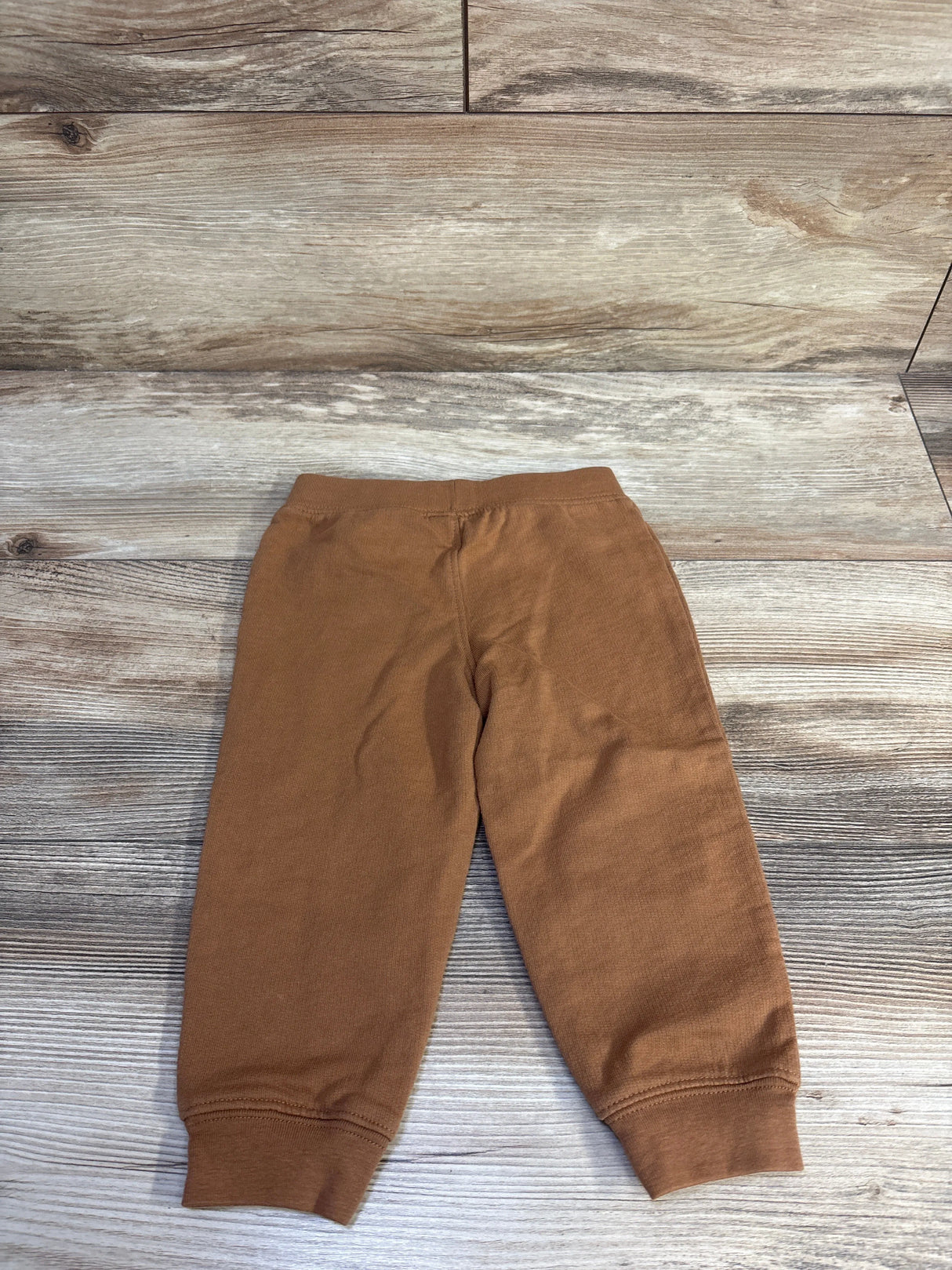 Top-down view of brown toddler joggers showing elastic waistband and cuffed ankles on light wood background, size 18 months