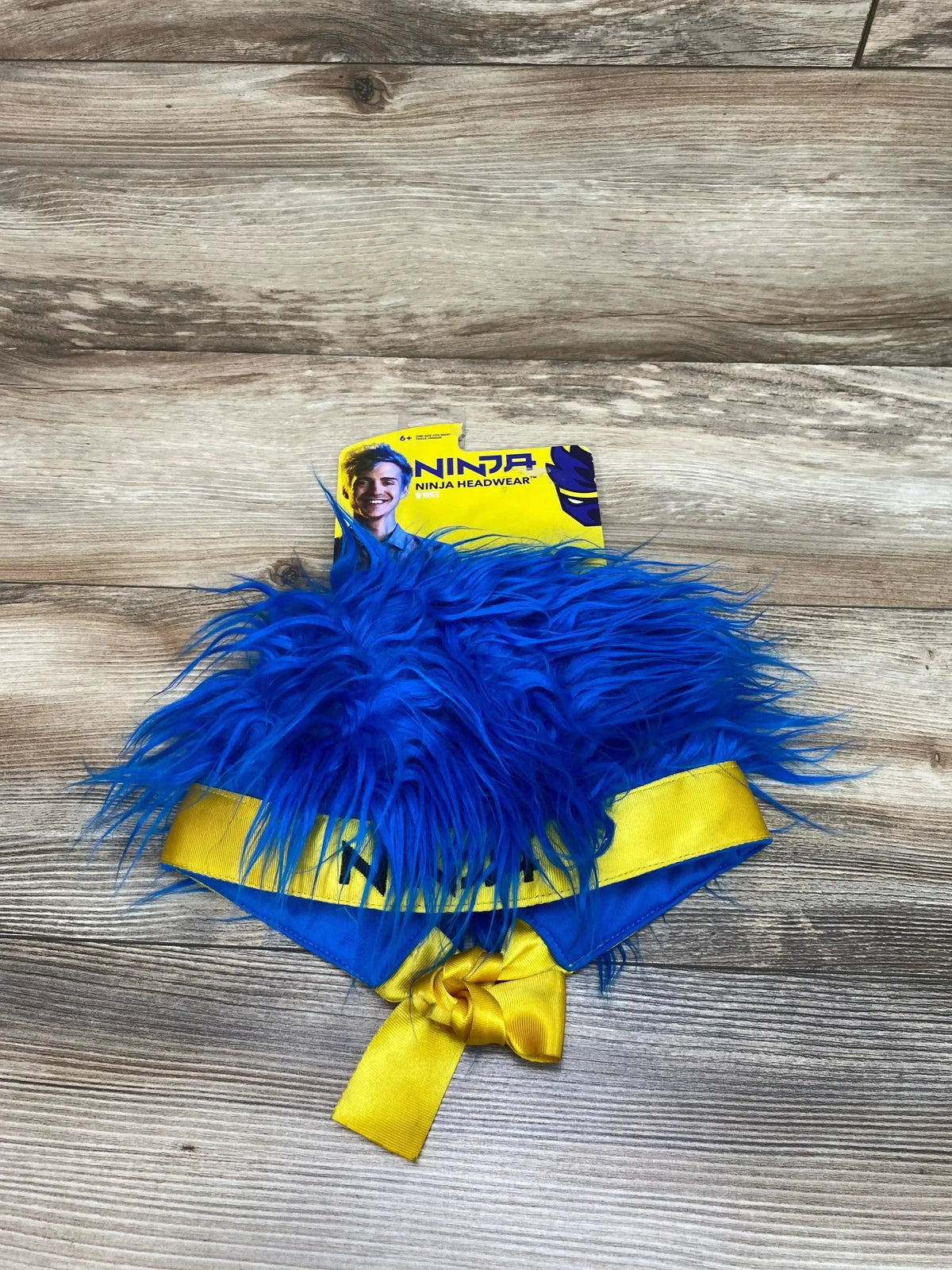 Front view of Ninja Blue Hair Headgear showcasing shaggy blue hair, yellow and blue trim, and tie on a wood grain surface