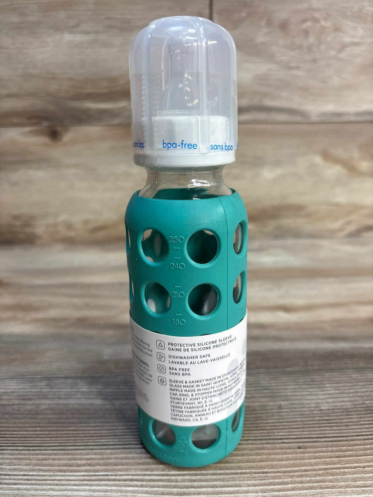 Glass baby bottle with teal silicone sleeve and BPA-free label, standing on wood surface