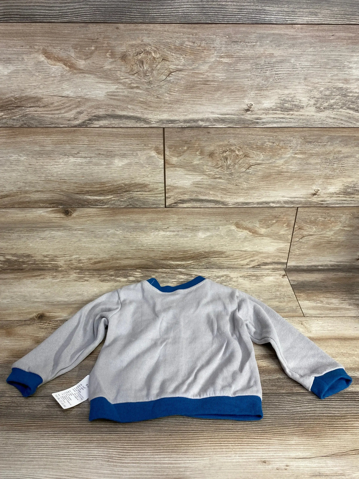 Back view of gray baby dinosaur button-front cardigan showcasing blue ribbed cuffs and hem with care label visible on wood floor