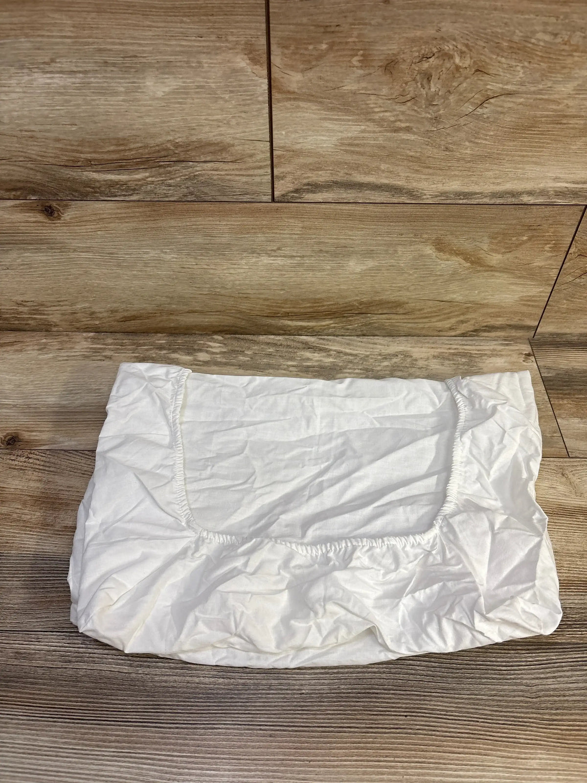Top-down view of white fitted bassinet sheet showcasing elasticized edges and rectangular mattress opening on wood floor