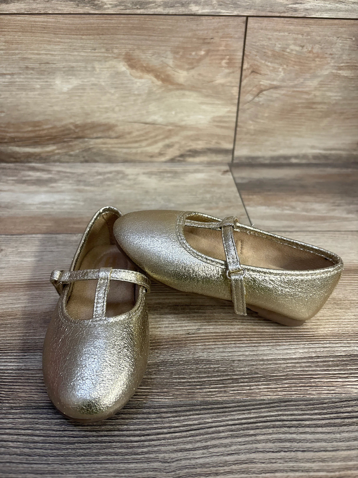 Angled view of Jumping Beans Susie T-strap ballet flats in metallic gold with textured upper and adjustable strap on wood