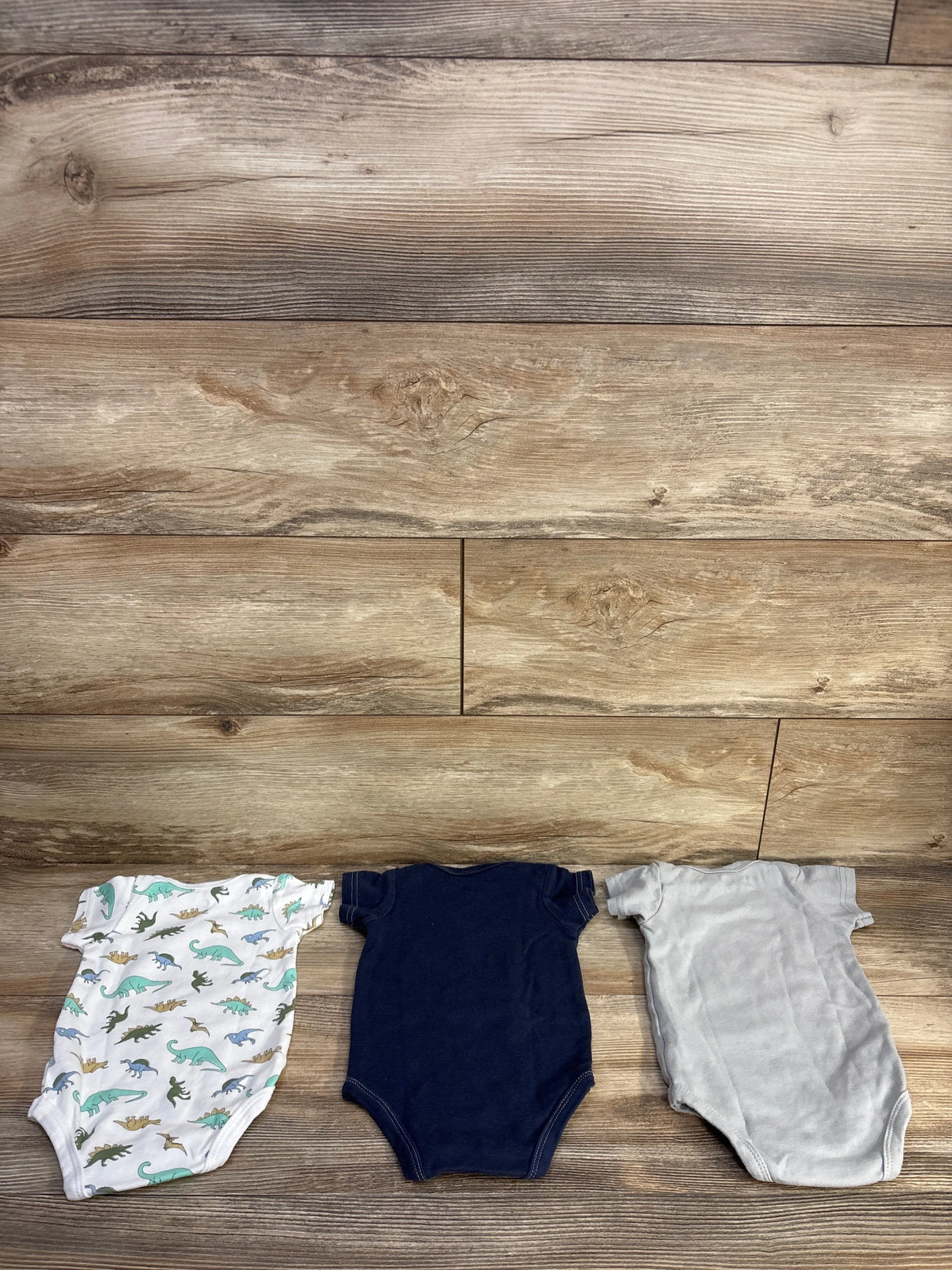 Flat lay of 3-pack 3-6M bodysuits showing dinosaur print, navy and gray solids with snap closures on wood