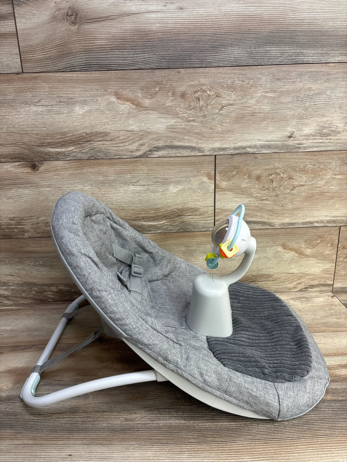 Angled view of Skip Hop Silver Lining Cloud upright floor seat showing gray fabric, built-in safety harness and attached toy bar on wood floor