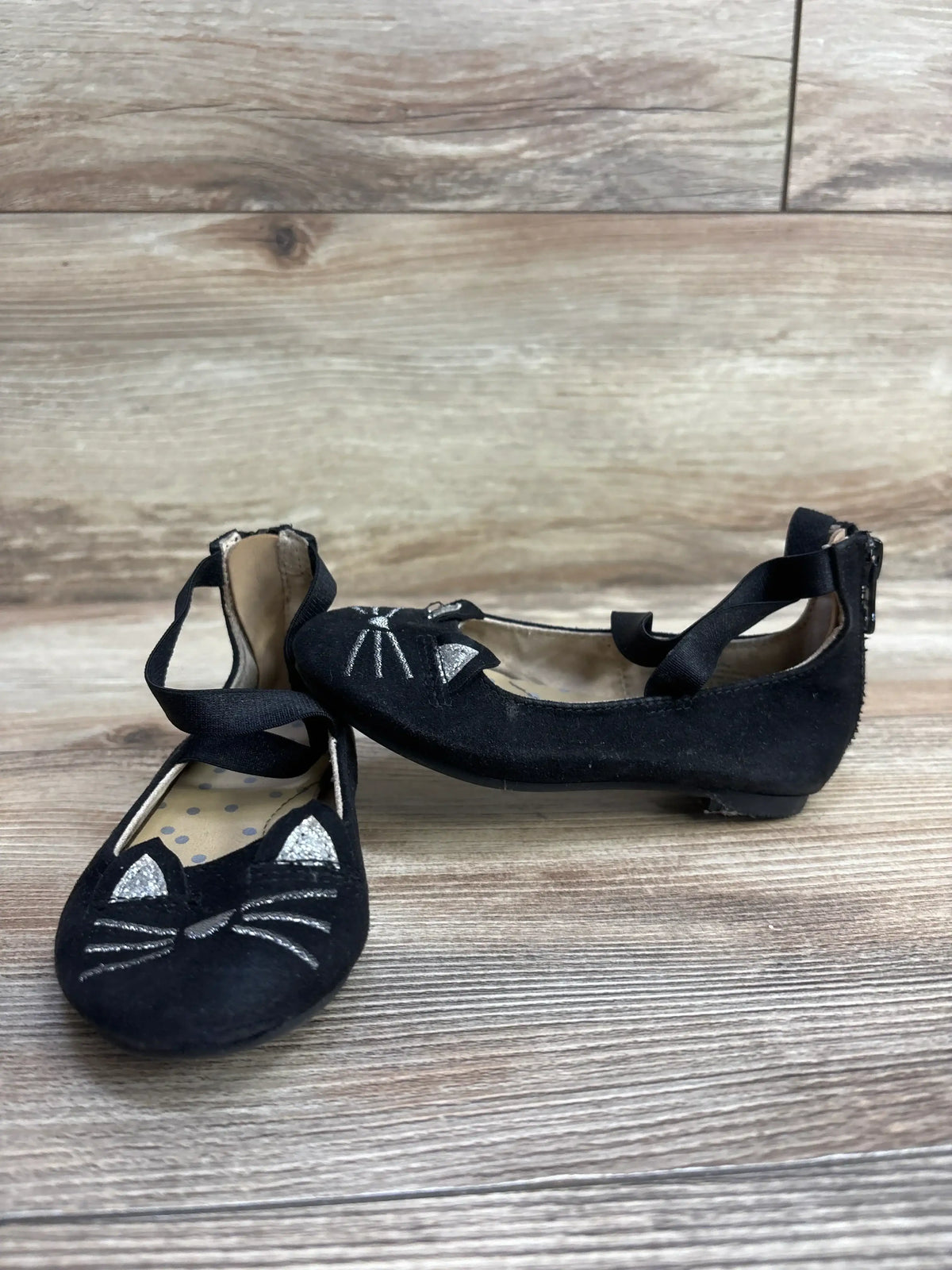 Black children's ballet flats with silver cat face embroidery and ankle straps on wood background