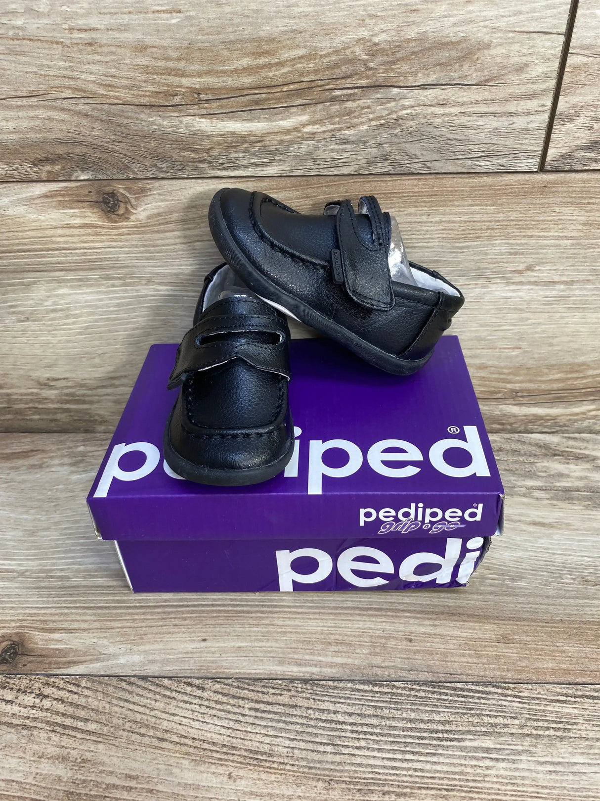 Angled front view of black toddler Pediped Charlie loafers with velcro strap on purple box and wood background