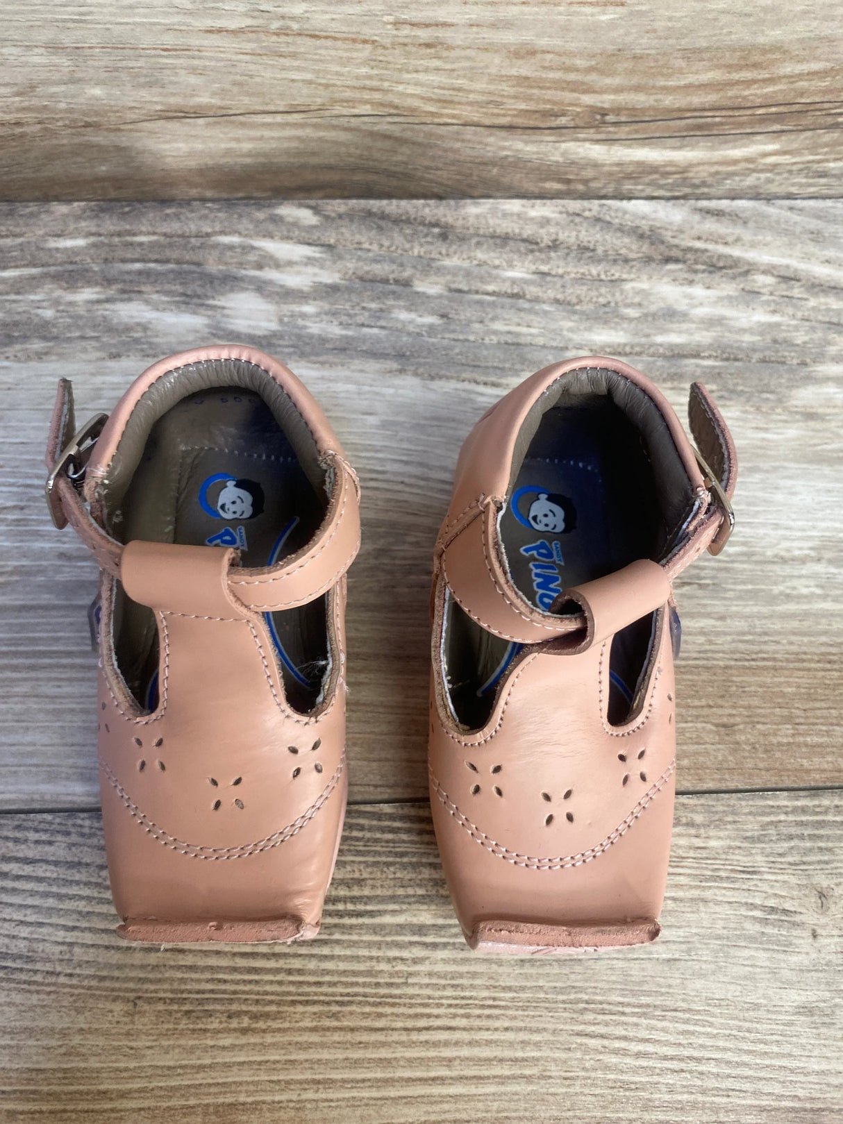 Pink leather baby shoes with strap and floral cutout design on wooden background