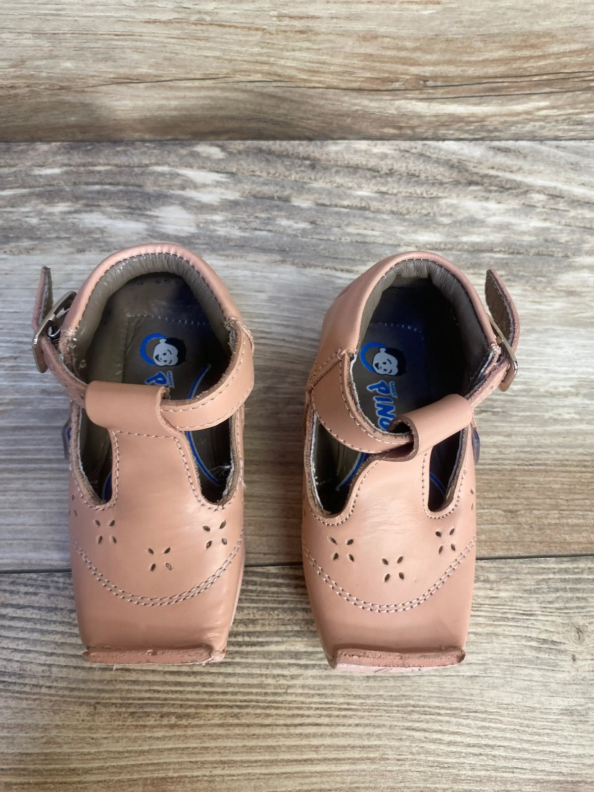 Pink leather baby shoes with strap and floral cutout design on wooden background