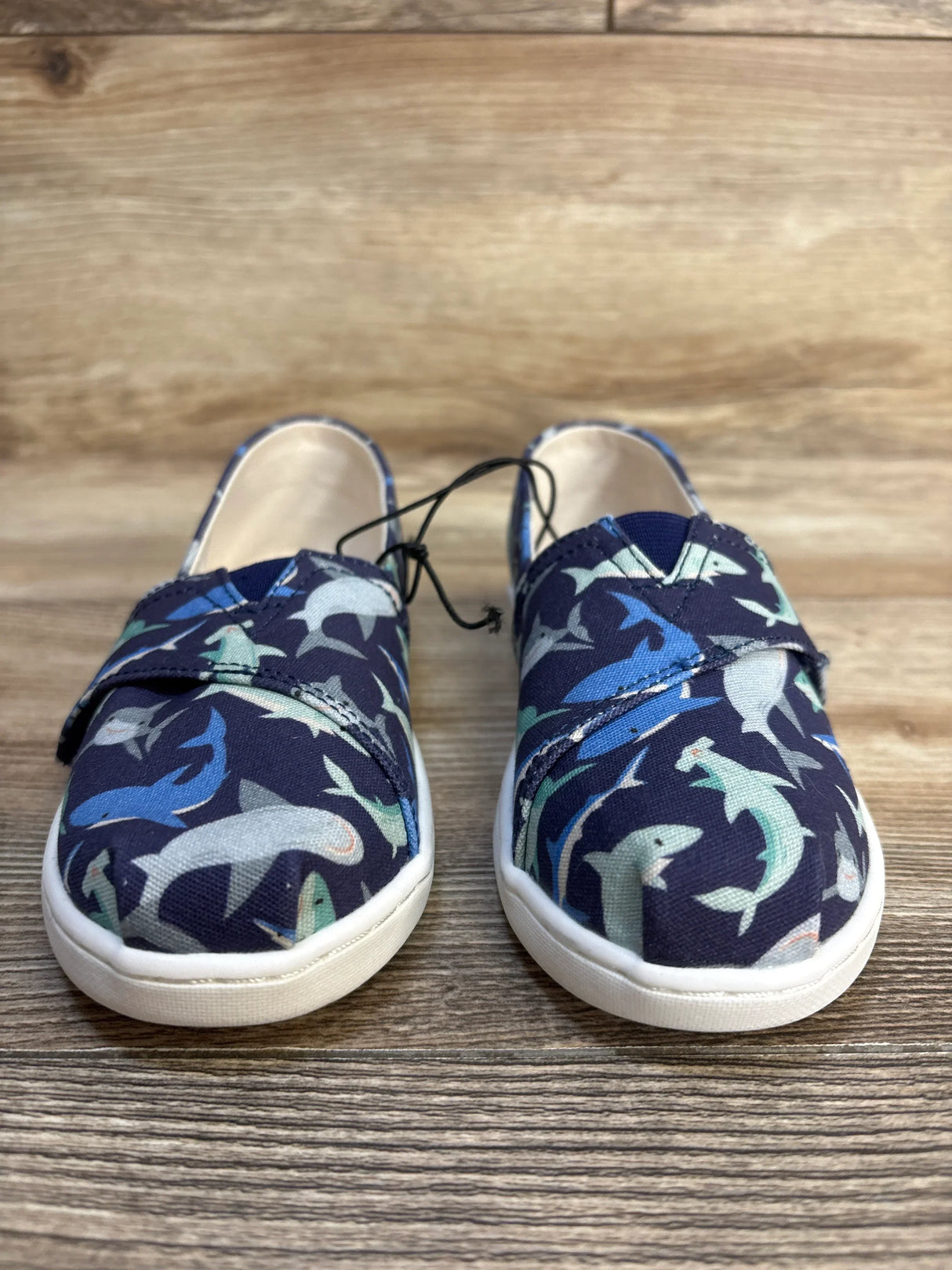 Kids' shark print slip-on sneakers with navy background, white soles, and black laces on wood floor