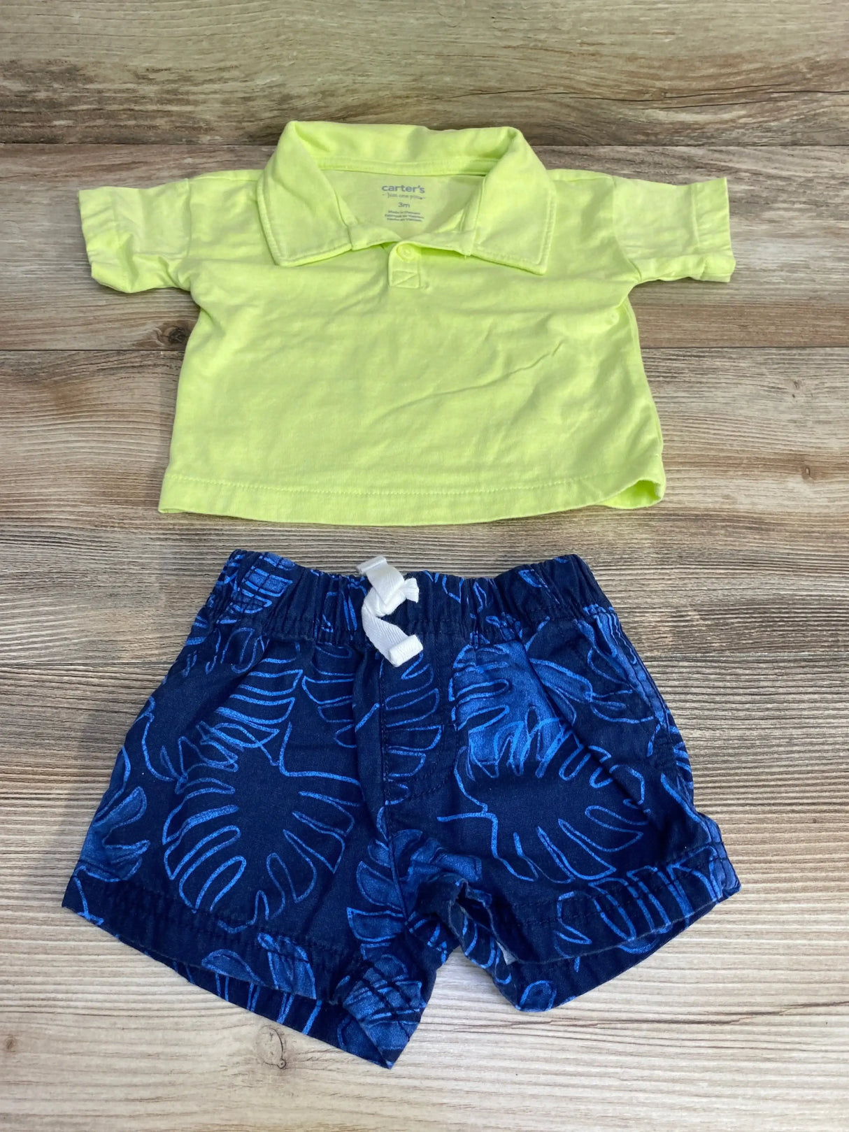 Flat lay of Just One You 2pc set with lime green polo shirt and navy blue palm leaf print drawstring shorts on wood plank floor