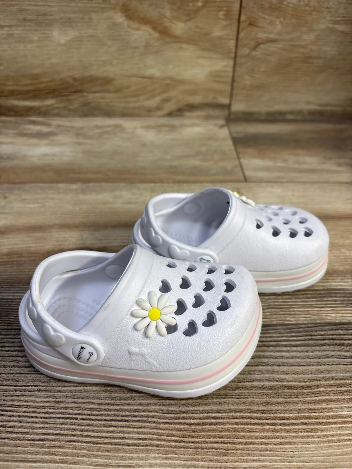 Angled side view of white Koala Kids toddler clogs showcasing heart-shaped cutouts, daisy charm, and pink-striped sole on wood floor background