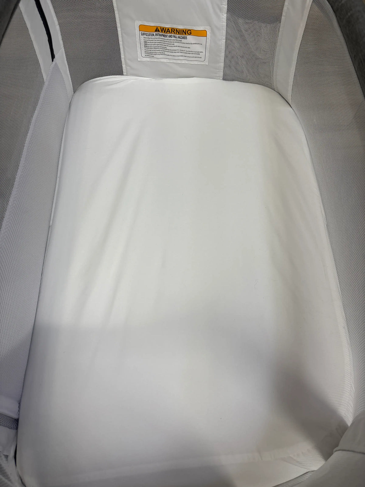 Interior view of Baby Delight Beside Me Dreamer bedside bassinet showcasing white mattress, mesh sides and warning label