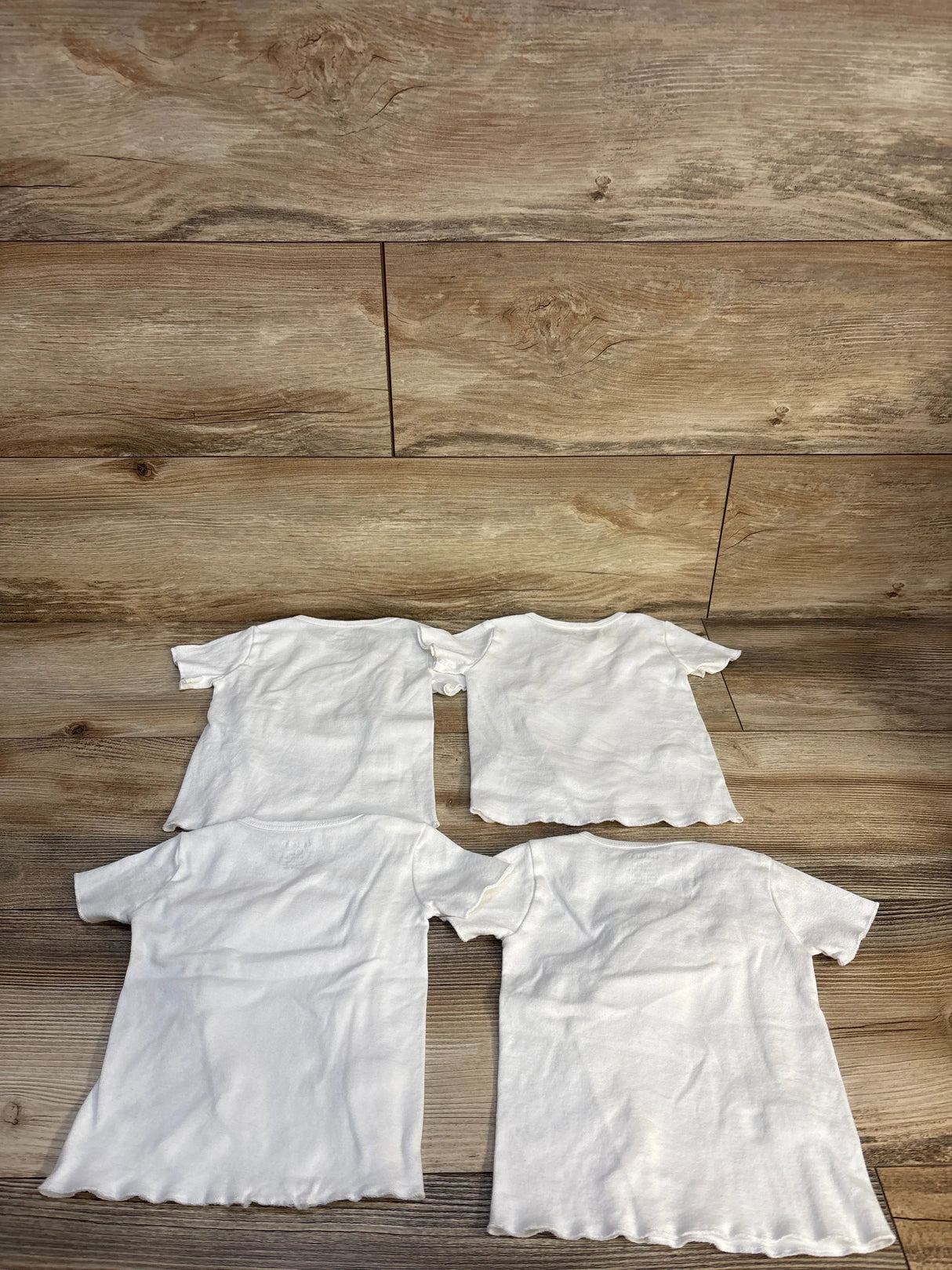 Flat lay of four Carter's white side-snap baby shirts (12M) showing short sleeves and scalloped hems on a wood background