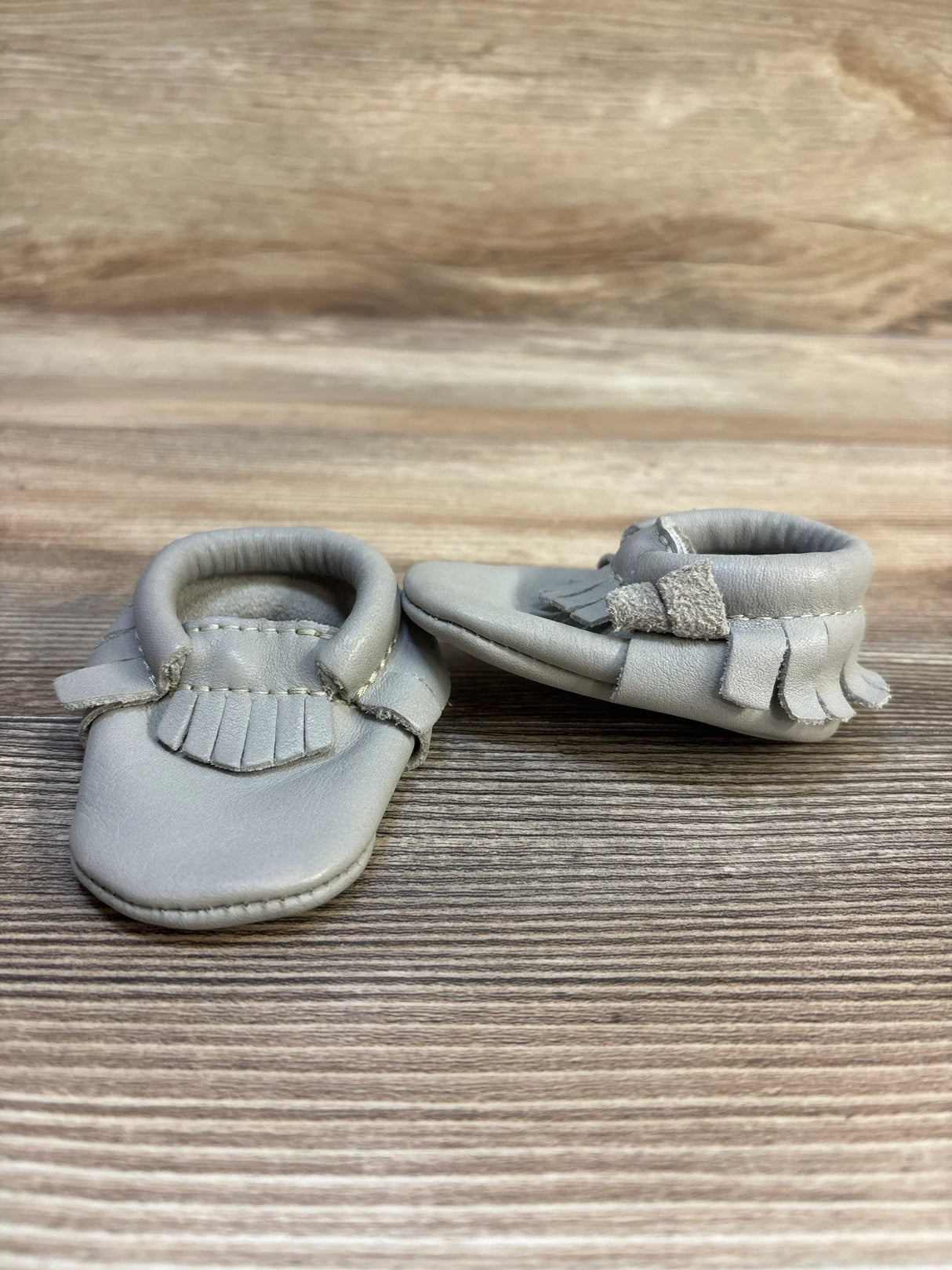 Front view of Freshly Picked Classic Mocc size 0C grey baby moccasins showing fringe detail on wooden surface, gently used
