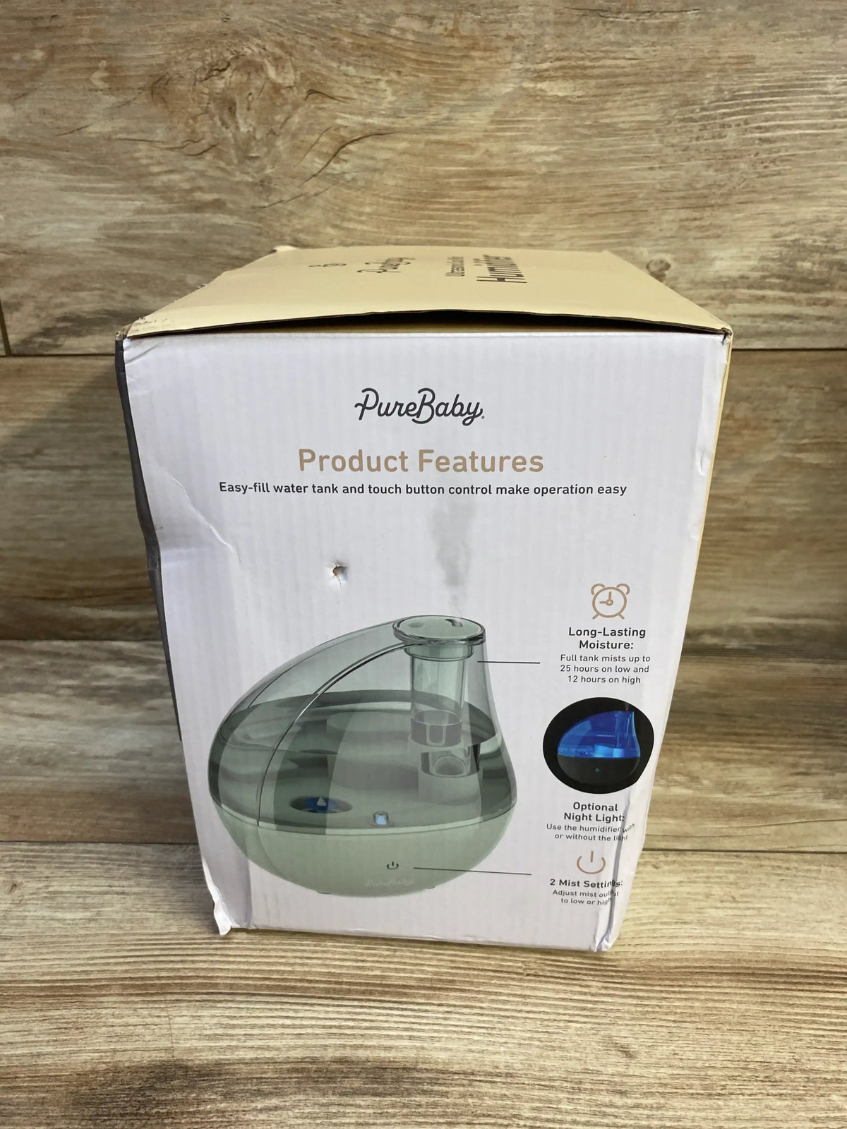 Front view of boxed PureBaby ultrasonic cool mist humidifier showcasing clear water tank and product features text, on wooden surface