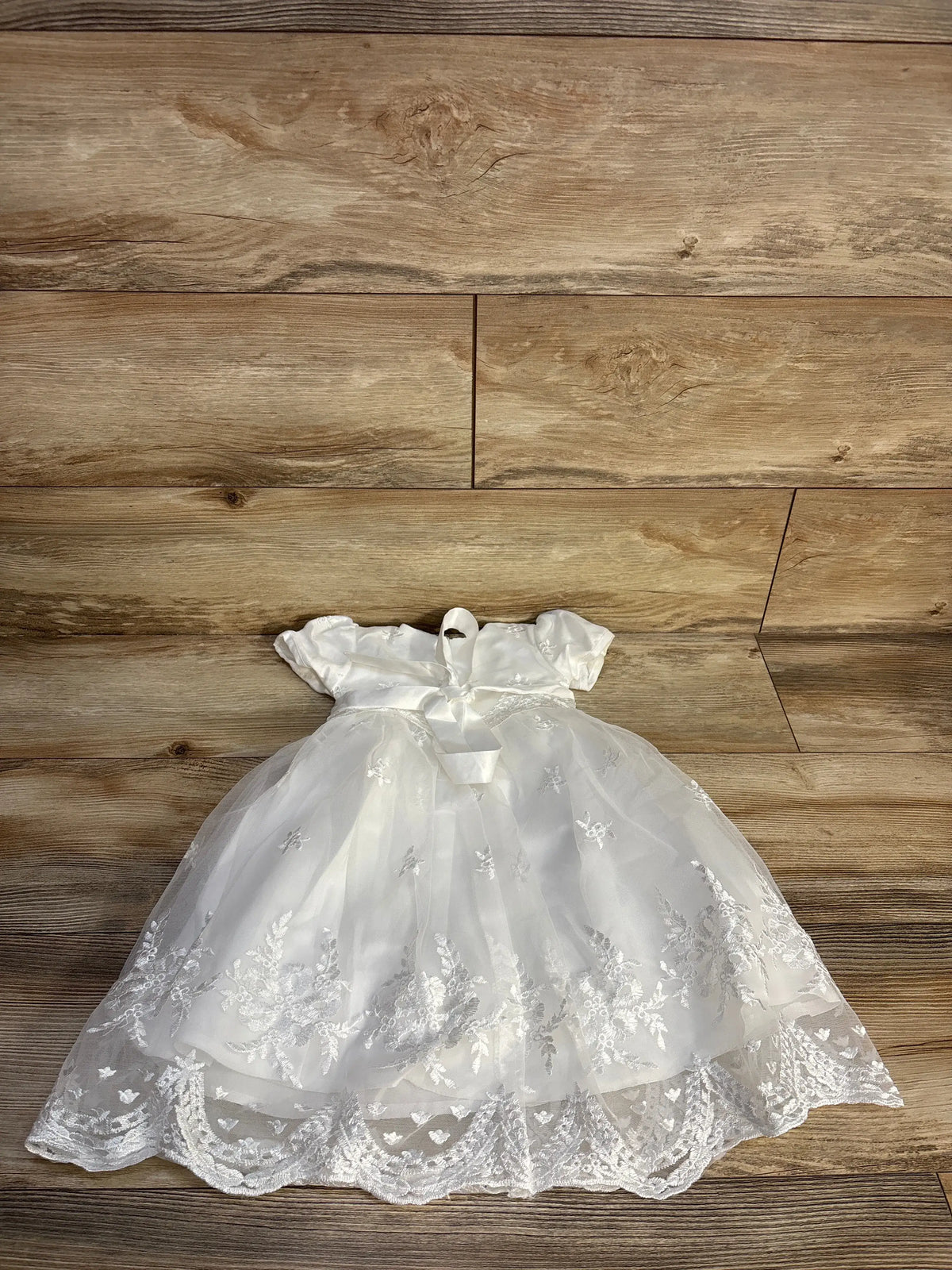 Front view of Glamulice christening and baptism dress showcasing white floral embroidery, cap sleeves, and bow on wood floor