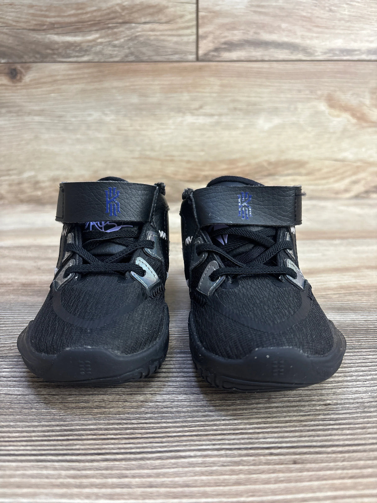 Black athletic sneakers with ankle straps and blue Kyrie logos on wood floor