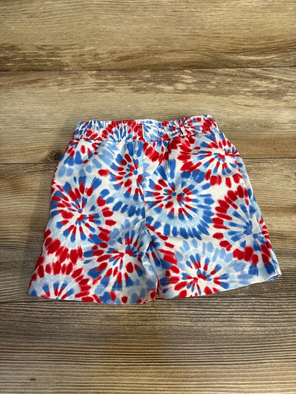 Front view of Jumping Beans 12M tie-dye swim trunks with red and blue spiral pattern and elastic waistband on wood floor