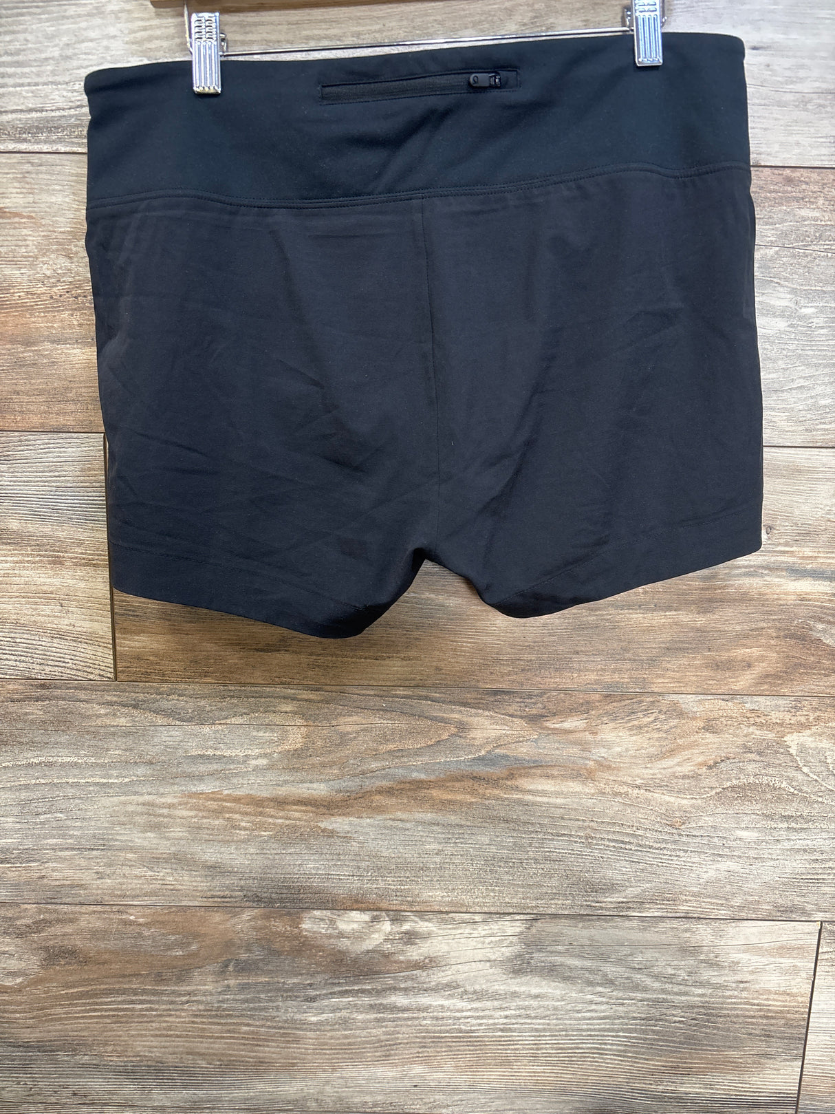 Front view of black GapFit maternity running shorts on hanger showing wide fold-over waistband with small zip pocket against wood plank background