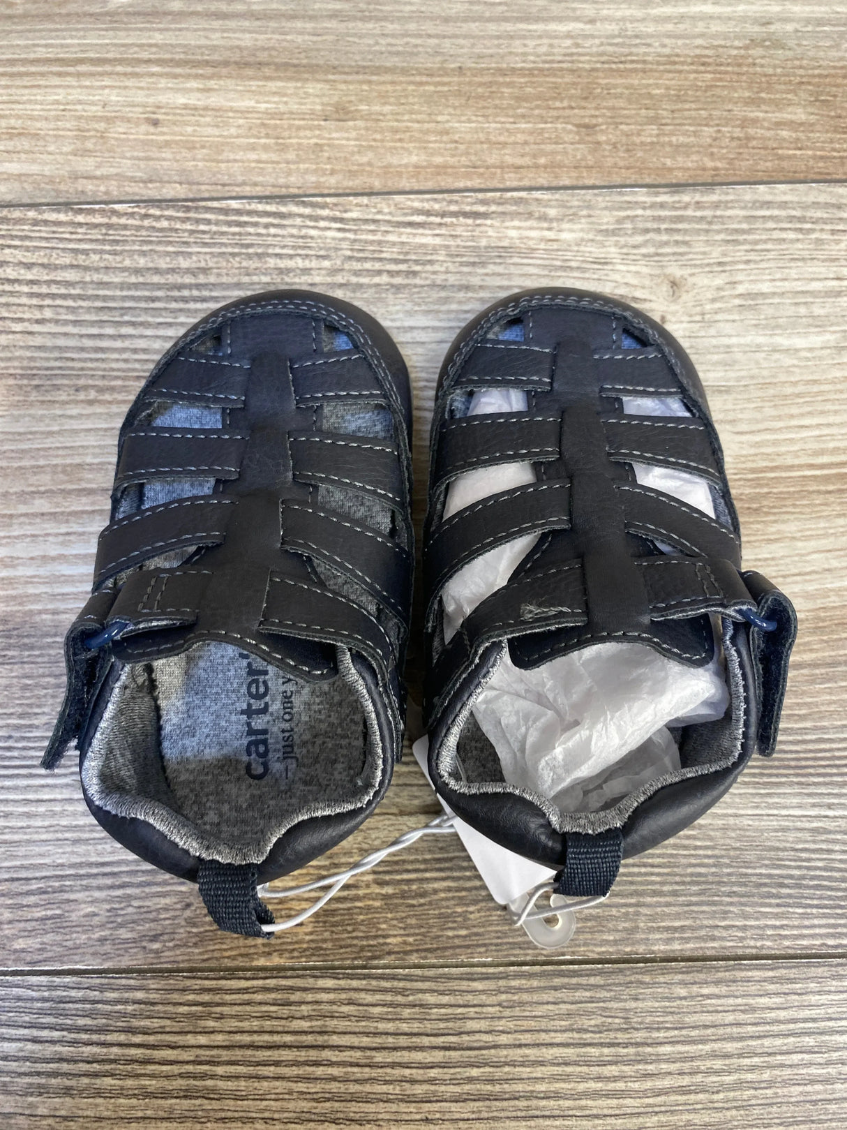 Top-down of Just One You Harrison fisherman sandals size 4/5c (12-18m) showing closed-toe navy straps and Velcro closures on wood