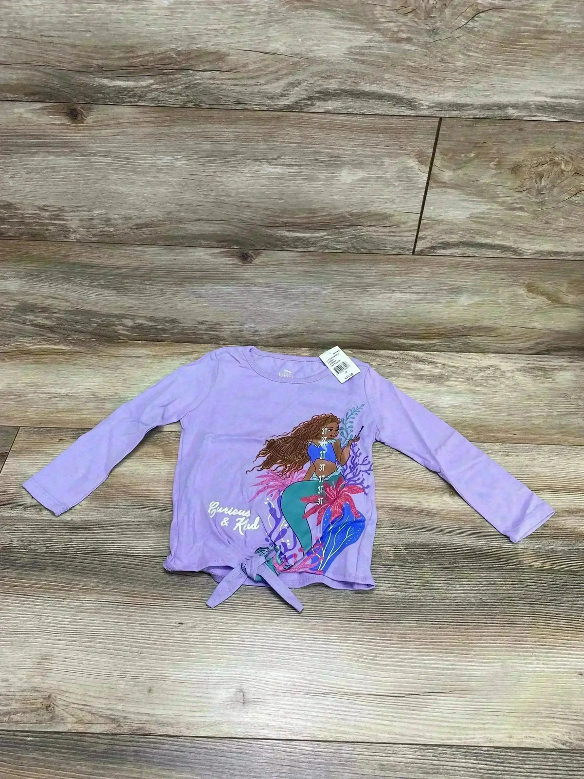 Front view of lavender long sleeve toddler top with mermaid print and knotted tie, size 3T tag visible on wood background