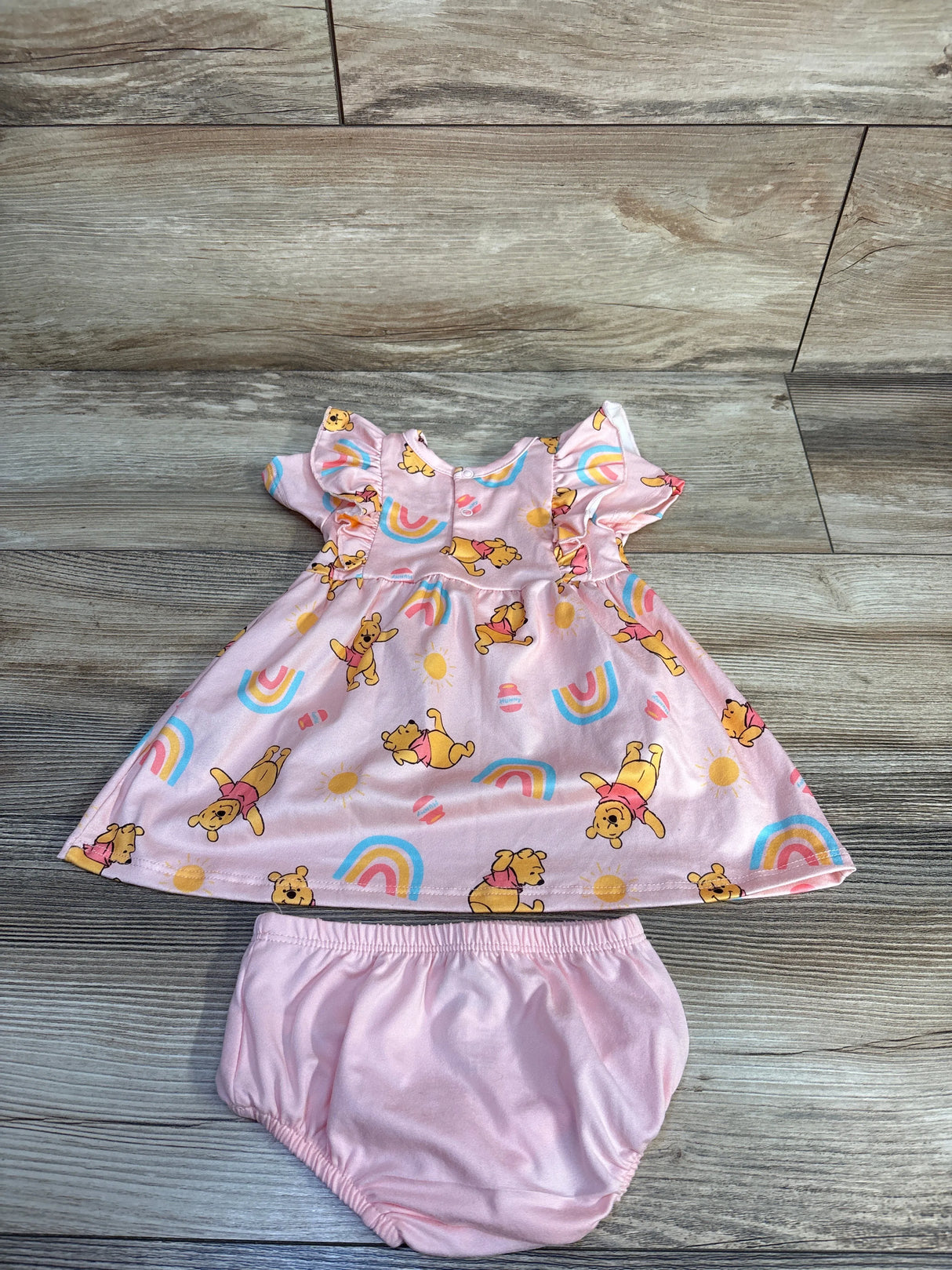 Flat lay of pink Disney Baby Winnie the Pooh dress and matching bloomers showing all-over Pooh and rainbow print on wood background
