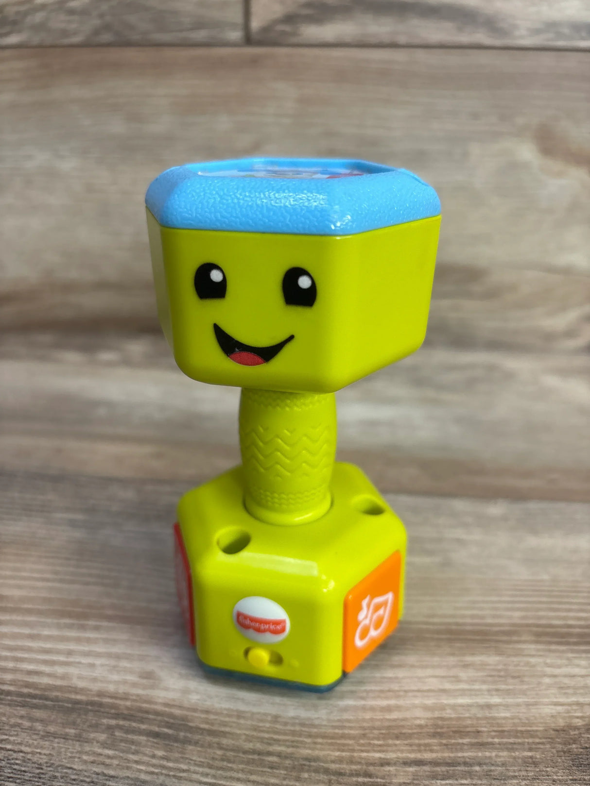 Angled front view of Fisher-Price Laugh & Learn Countin' Reps green dumbbell rattle showcasing smiling face, textured grip, and orange music button on a wood surface background